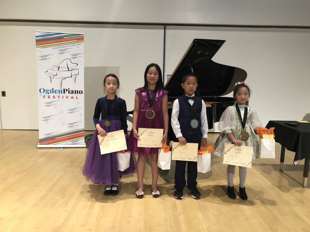 Elementary Division Winners with certificates and prize bags in front of Steinway Grand Piano and Ogden Piano Festival banner. Left to Right: Abigail Yang, Honorable Mention; Annie Jia, 3rd Place; Maxwell Ji, 2nd Place; Natalie Sun, 1st Place.