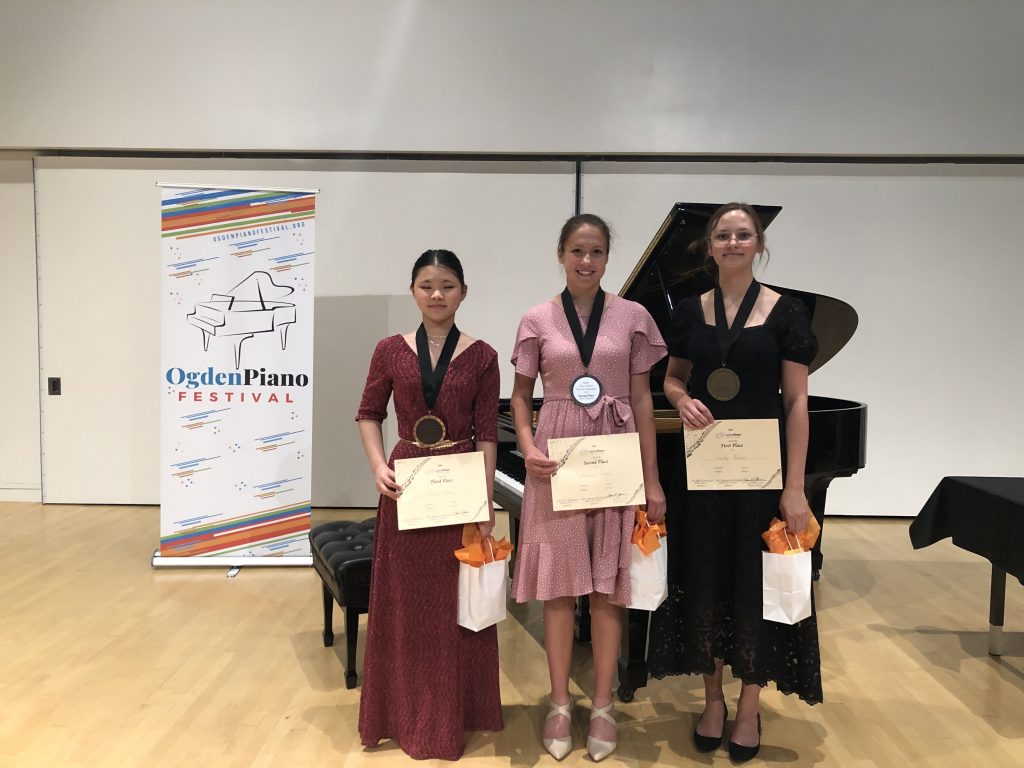 Senior division winners L to R: Aimee Lam, 3rd; Katherine Leach, 2nd; Emily Runov, 3rd. Standing with certificates and gift bags in front of piano and Ogden Piano Festival Banner.