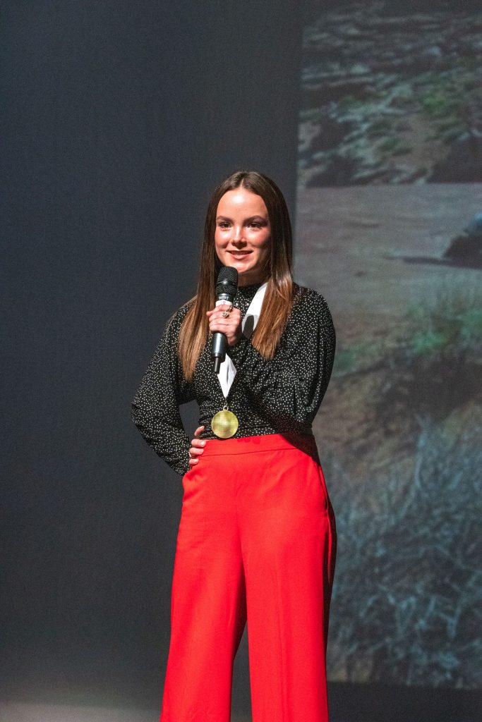 Genevieve Fullmer, Guest Speaker stands on stage. There is a picture of desert on the projector screen behind her.