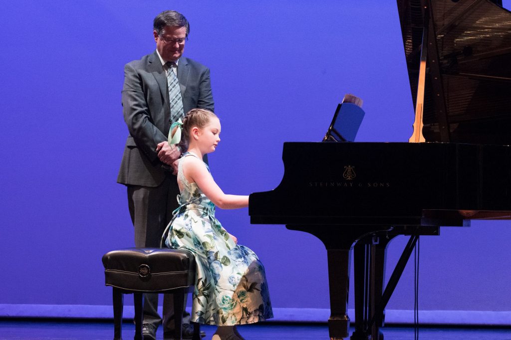 Elle Hirst plays the piano with her father standing behind her. 